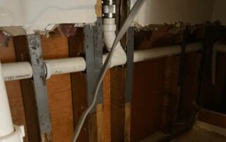 A close-up view of exposed PVC and copper plumbing pipes within a damaged wall, showing wooden studs and drywall removed in sections. A new silver coupling connects a copper pipe at the top, leading down to a white PVC pipe.