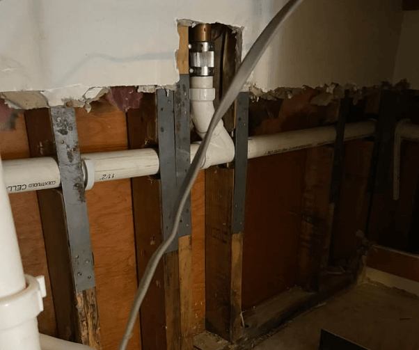 A close-up view of exposed PVC and copper plumbing pipes within a damaged wall, showing wooden studs and drywall removed in sections. A new silver coupling connects a copper pipe at the top, leading down to a white PVC pipe.