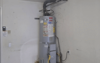 Vertical residential water heater tank, likely gas or electric, installed in a garage or utility corner with dirty white walls, showing power cord plugged into an outlet and exhaust venting near the ceiling.