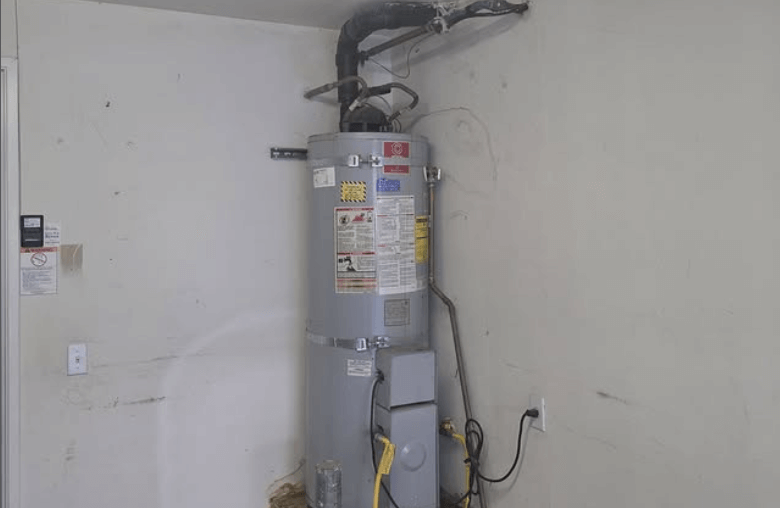 Vertical residential water heater tank, likely gas or electric, installed in a garage or utility corner with dirty white walls, showing power cord plugged into an outlet and exhaust venting near the ceiling.