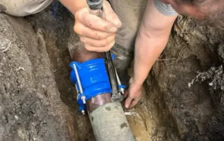 Plumber repairing an underground burst water pipe with a blue clamp in a muddy trench