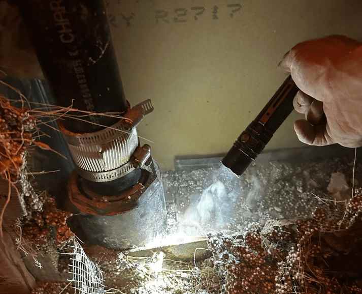 Plumber inspecting a leaking cast iron waste pipe with a flashlight in a crawl space