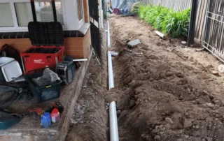 Excavated trench beside a house showing new PVC drain pipes being installed during a sewer line replacement project