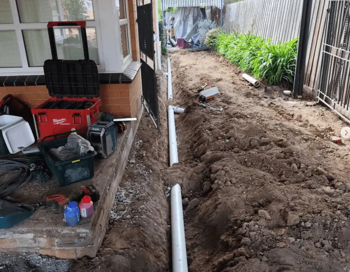 Excavated trench beside a house showing new PVC drain pipes being installed during a sewer line replacement project