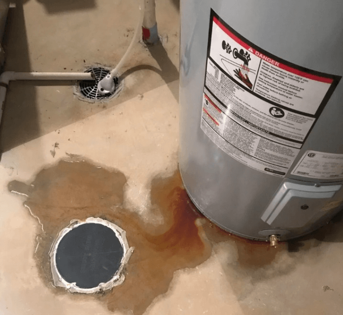 leaking-water-heater-on-basement-floor Water heater leaking rusty water onto a basement floor near a floor drain, showing signs of tank corrosion and water damage.