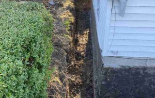 A long, narrow trench dug in the ground alongside a white house, indicating a traditional excavation for sewer line repair or replacement. Green bushes are visible on the left.