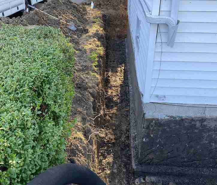 A long, narrow trench dug in the ground alongside a white house, indicating a traditional excavation for sewer line repair or replacement. Green bushes are visible on the left.
