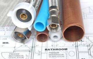 Close-up of copper, PEX, and flexible plumbing pipes with fittings placed on a bathroom blueprint, showing materials used in home piping systems