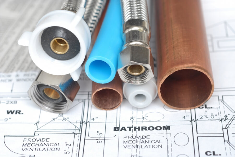 Close-up of copper, PEX, and flexible plumbing pipes with fittings placed on a bathroom blueprint, showing materials used in home piping systems