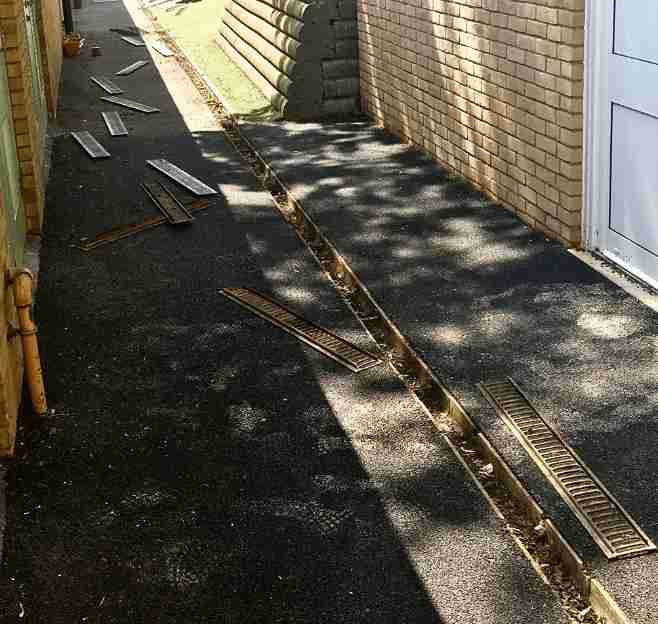 sewer-drain-cover-repair-along-walkway Drainage channel with metal covers removed along a paved walkway beside a brick wall, showing an open trench for sewer or surface water repair work.
